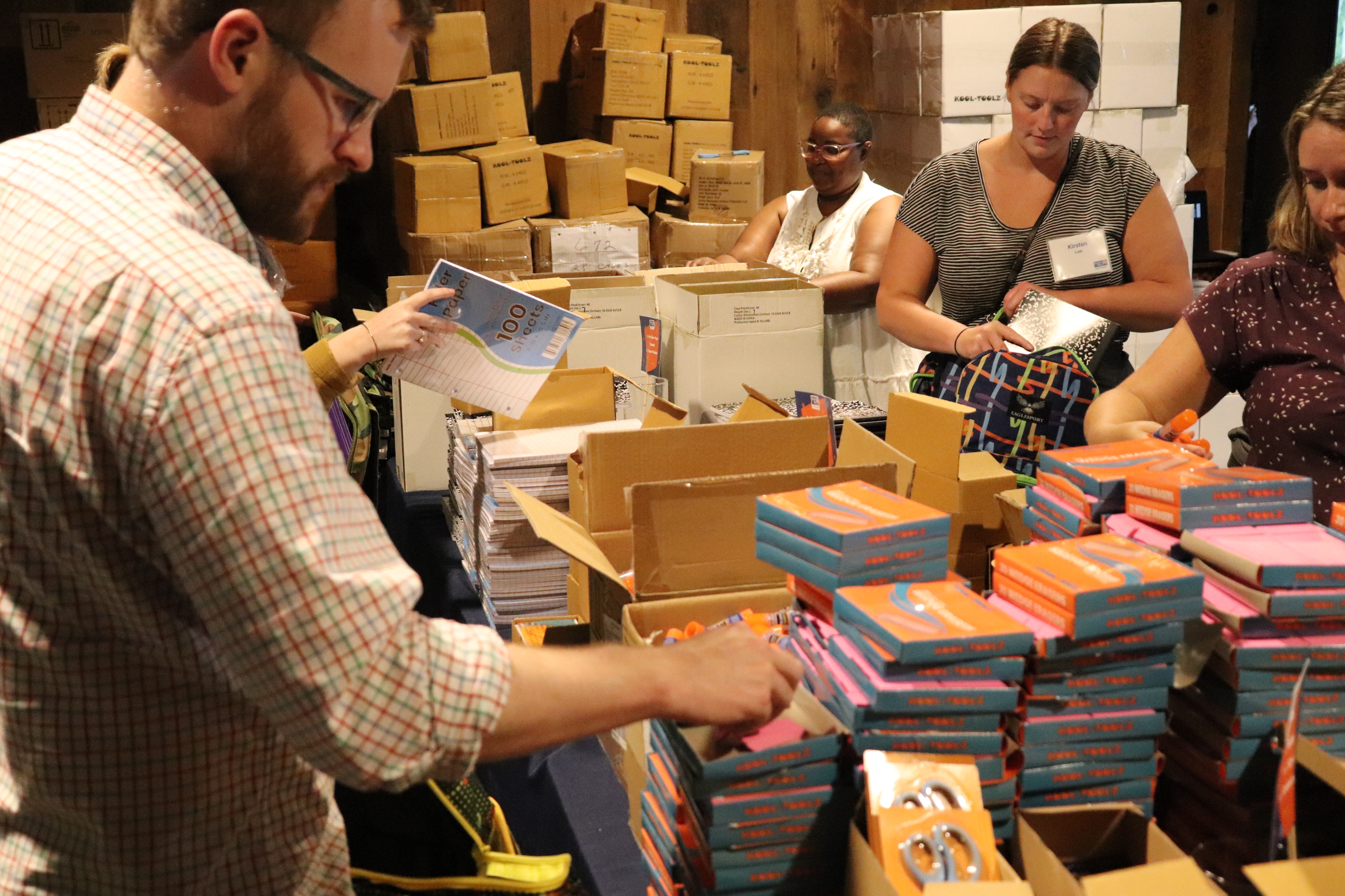 "Multiple volunteers stand around a pile of carboard boxes, books, and other school supplies."