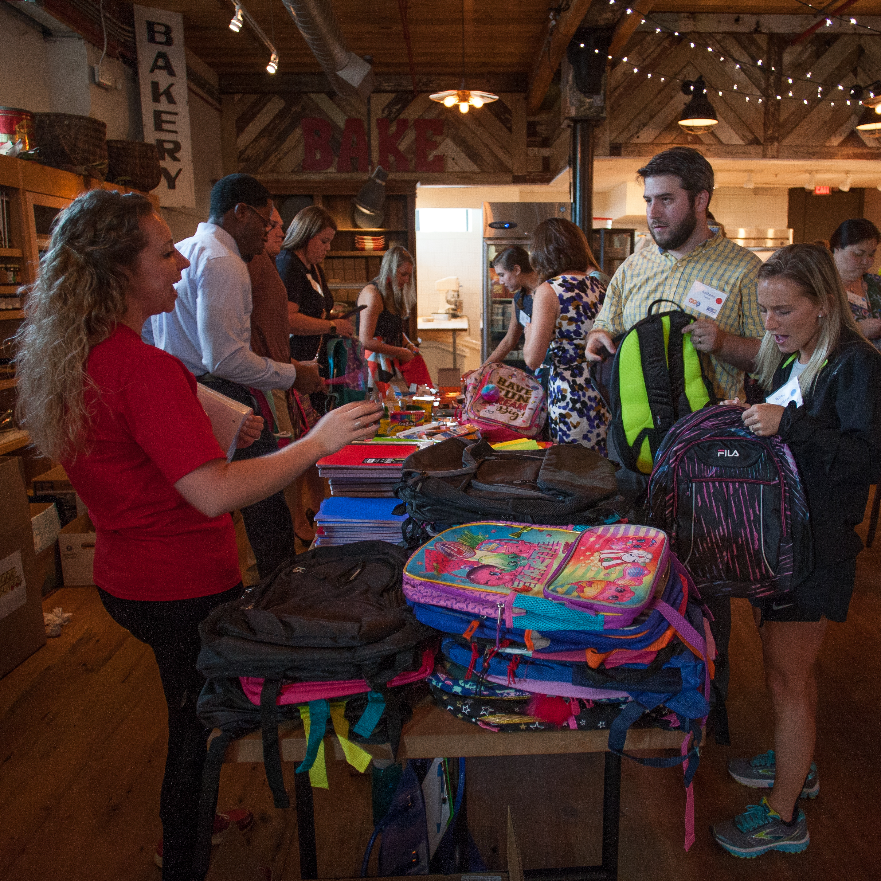 "Volunteers stand and chat on two sides of a long table. The table is covered in childrens' backpacks and school supplies, being stuffed by the volunteers."