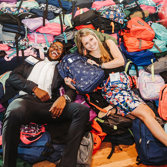 "Two volunteers lay smiling on top of a large pile of backpacks."