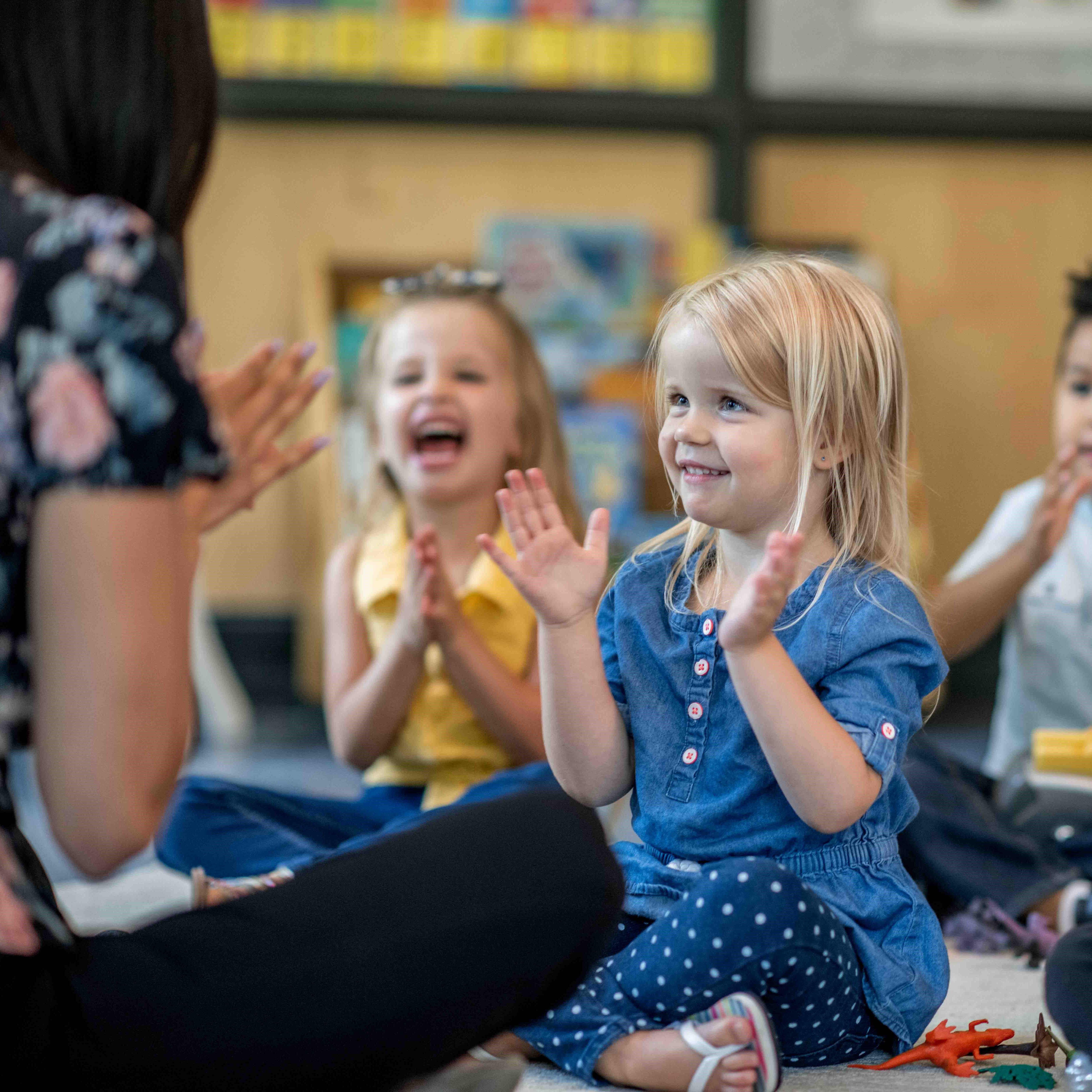 A child is seen sitting on the floor clapping with her teacher and class.