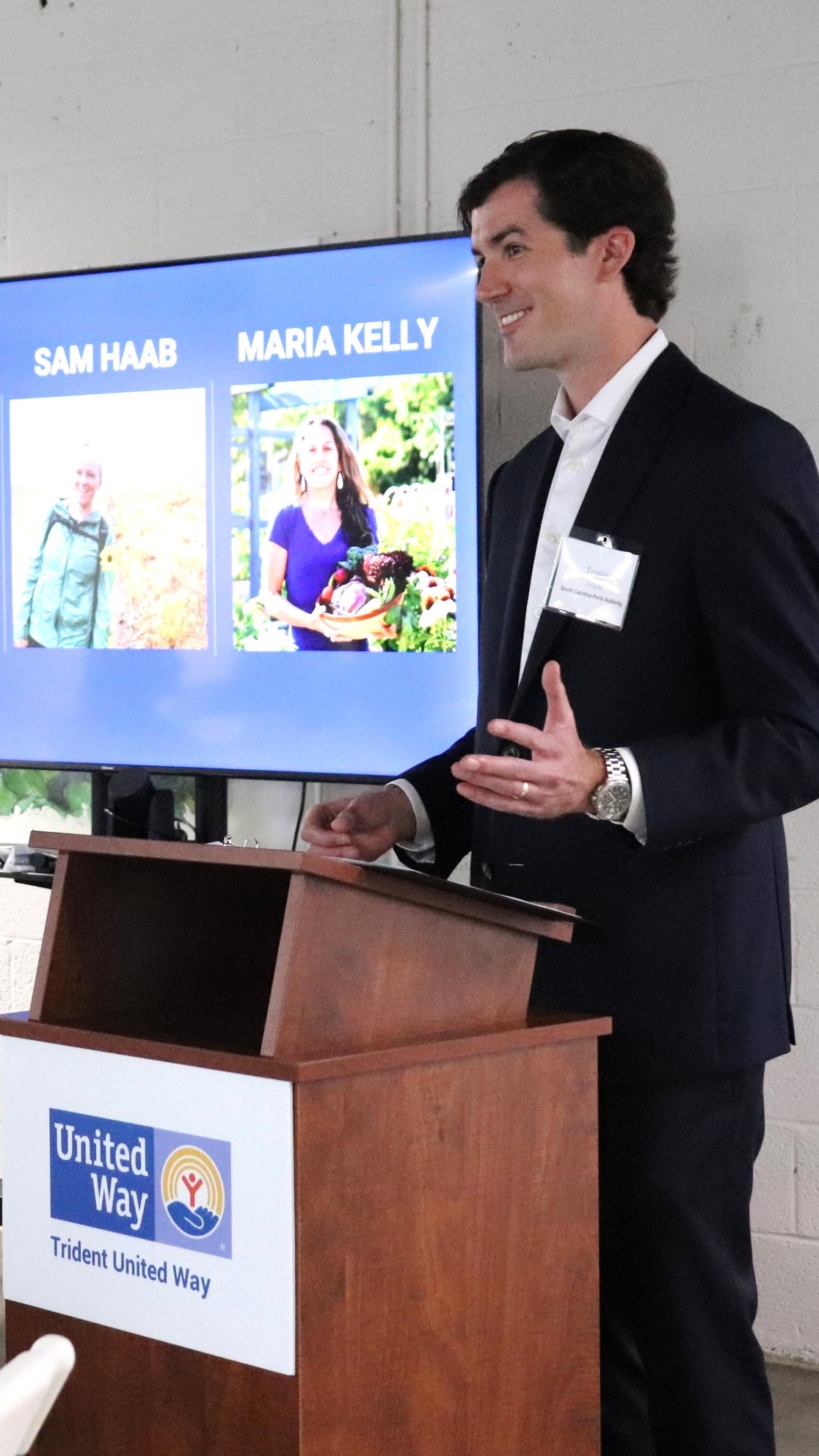 A speaker stands in front of a podium presenting information in a suit. He is smiling