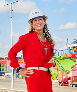 Barbara is wearing a red outfit and hard hat, standing in front of a blue sky and a maritime port.