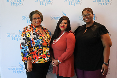 Three Black women smiling, standing against a step and repeat with the Sharing Hope SC logo.