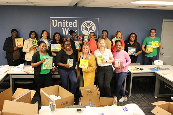 large group of people, holding books and smiling for the camera. They are standing in front of a United Way sign and there are boxes around them.