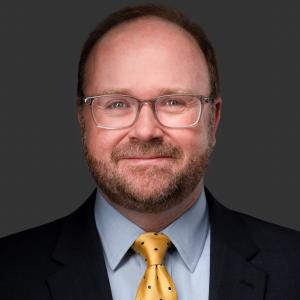 DJ Hampton is wearing glasses, a yellow tie with black dots, a light blue button up and black suit jacket. He is smiling at the camera.