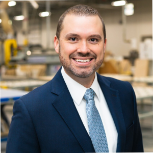 David Stasiukaitis is has short dark hair and short facial hair, wears a blue tie, white button up shirt and dark blue suit jacket. He is smiling at the camera