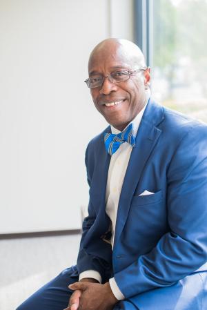 Herbert Drayton is wearing glasses, a bright blue bowtie, white button up shirt, blue suit jacket and blue pants. He is smiling looking at the camera.