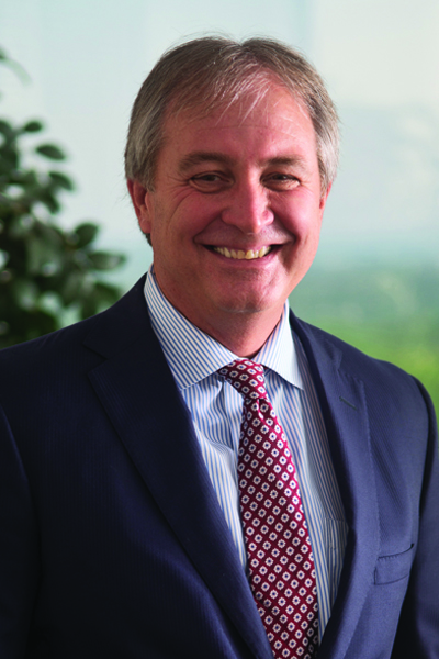 Tim Grow has short hair, is wearing a blue striped shirt, navy blue blazer and a red tie with a blue and white flower pattern. He is looking at the camera, smiling.