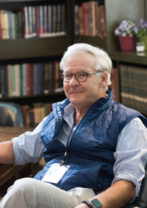 Walter has short white hair and wears glasses. He has on a blue vest, and blue stripped button up shirt. He is looking at the camera smiling.