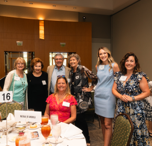 A group of attendees standing together smiling at the camera. One woman is seated at the table.