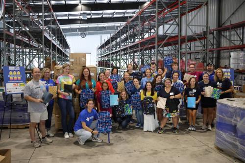 A group of 30 volunteers pose in a warehouse setting holding backpacks and school supplies