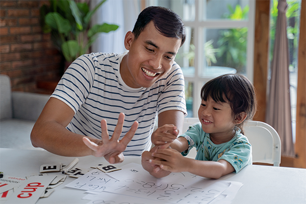 A parent is teaching his daughter how to count. He is holding up four fingers and smiling, She is watching him smiling. There are flashcards on the table with words and letters, and papers with math problems