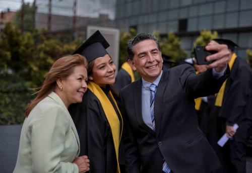 Photo of a family taking a photo at their daughter's graduation. Mom is on the left, daughter in the middle wearing cap and gown and father on the right holding a camera to take a selfie 