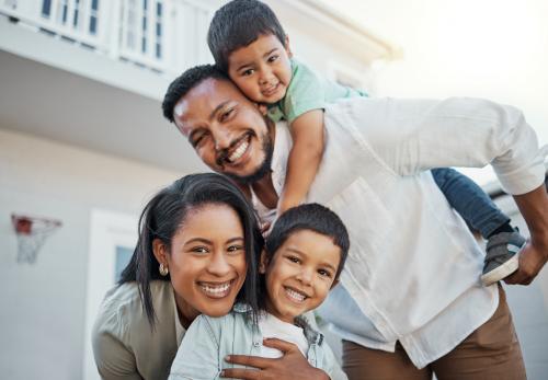 A family of four, two adults and two children, smile at the camera while outside in front of their home