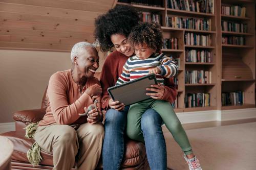 An older woman sits in a chair, younger woman sits on arm and child sits in her lap. They are all looking at an ipad and smiling