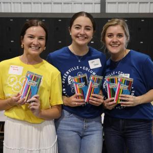 Three people hold up pencils while volunteering to help pack school supplies.