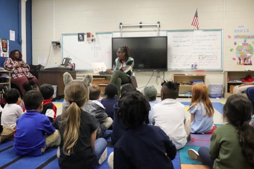 A woman sits on a chair in a classroom, all the children are sitting in front of her on the floor. She is in the middle of reading a book aloud to them with excitement