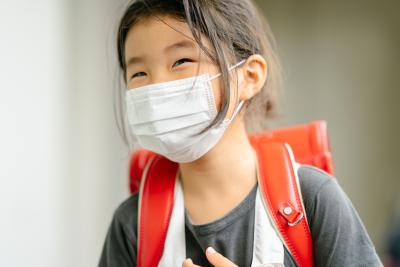 A child wearing a facemask smiles with a backpack on.