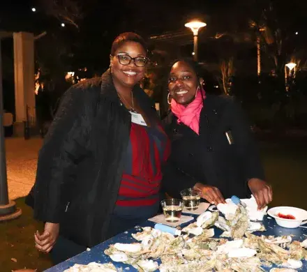 Two women are standing and smiling at the camera at a table with oysters