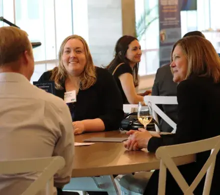 Two women are sitting at a table, both are smiling and looking at the man sitting at the table with them. There are groups of tables behind them in the room