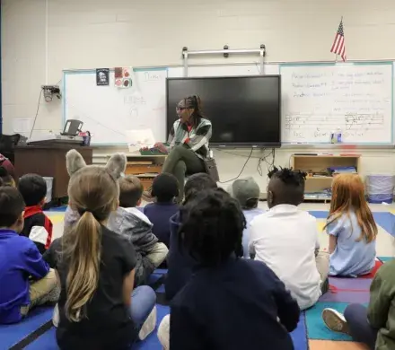 A woman sits on a chair in a classroom, all the children are sitting in front of her on the floor. She is in the middle of reading a book aloud to them with excitement