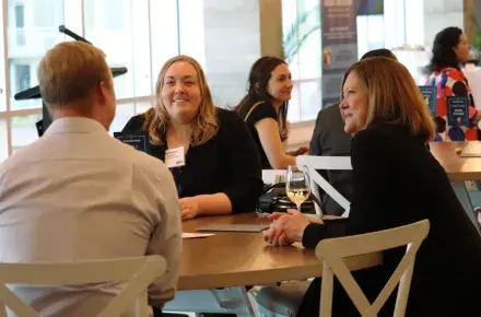 Two women are sitting at a table, both are smiling and looking at the man sitting at the table with them. There are groups of tables behind them in the room