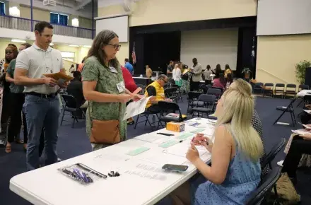 Photo of a woman standing at a folding table, holding play money and a money pouch. She is talking to two people sitting on the otherside of the folding table.