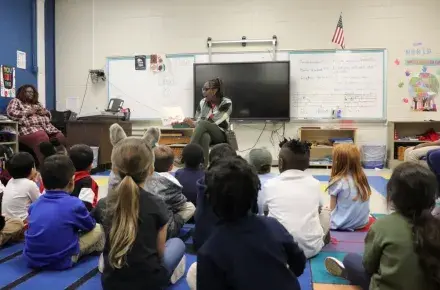 A woman sits on a chair in a classroom, all the children are sitting in front of her on the floor. She is in the middle of reading a book aloud to them with excitement