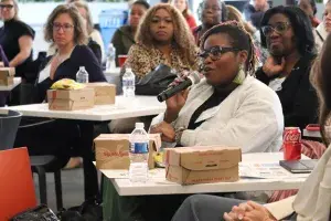 A group of women sitting at lunch tables for a Women United lunch and learn event. One woman holds the microphone and is speaking