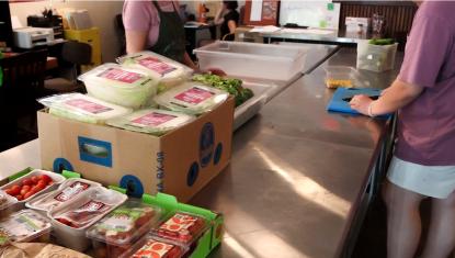 Boxes of vegetables on a metal table, two individuals cutting vegetables on cutting boards