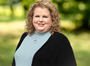 Headshot photo of Andrea Boccucci in front of a tree. She is wearing a light blue turtle neck with a black cardigan. Andrea has short curly blonde hair. 