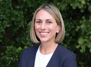 Photo of Devon Wade outside in front of a tree with green leaves. She is smiling, has shoulder length hair, wearing a white top and a navy blue blazer