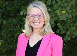 Julie Frye is standing outside in front of a tree with green leaves. She is smiling, wearing glasses, black top and a pink blazer.