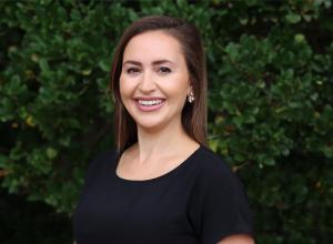 Sarah Makuta is posing outside in front of a tree with green leaves wearing a black short sleeve top.