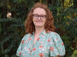 Kimberly wears glasses and a light blue floral dress. She has shoulder length red curly hair.