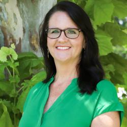 Photo of Amy Maciariello outside in front of a tree. She is wearing a kelly green v-neck blouse and black glasses. Amy has shoulder length straight dark hair.