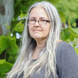 Photo of Angela Boltz outside in front of a tree. She is wearing a dark gray long sleeve shirt and thin black glasses. Angela has long wavy gray hair.