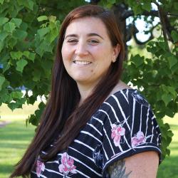 Photo of Ashley Morgan outside in front of a tree. She is wearing a black and white striped shirt with pink flowers. Ashley has long straight dark brown hair.