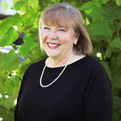 Photo of Cathy Easley outside in front of a tree. She is wearing a black blouse with a pearl necklace. Cathy has short brown hair with bangs. 