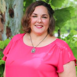 Photo of Christy Boudolf outside in front of a tree. She is wearing a dark pink blouse with a silver pendant necklace. Christy has short curly brown hair. 