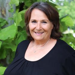 Photo of Debbie Mann outside in front of a tree. She is wearing a black long sleeve blouse. Debbie has short dark brown hair.