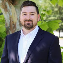 Photo of Joey Current outside in front of a tree. He is wearing a navy suit jacket with a white button down shirt. Joey has short dark brown hair and a beard. 