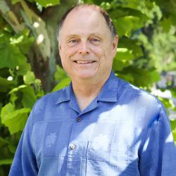 Photo of John Boyle outside in front of a tree. He is smiling wearing a blue button down shirt. John has short brown hair. 