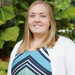 Photo of Katherine Torbush outside in front of a tree. She is wearing a blue chevron patterned blouse with a white cardigan. Katherine has shoulder length blonde hair. 