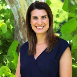 Photo of Katie Reams outside in front of a tree. She is wearing a navy, sleeveless, v-neck blouse. Katie has shoulder length brown hair. 