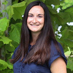 Photo of Kelli Morse outside in front of a tree. She is wearing a navy blue blouse. Kelli has straight long dark brown hair. 