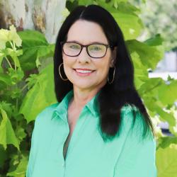 Photo of Louise Lawton outside in front of a tree. She is wearing a mint green button down shirt, black glasses and gold hoop earrings. Louise has shoulder length straight dark hair.