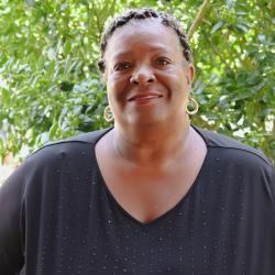 Photo of Melrose Smith outside in front of a tree. She is wearing a black shirt and gold hoop earrings. Melrose has braided dark hair.