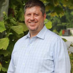 Photo of Michael Kirchner outside in front of a tree. He is wearing a light blue plaid button down shirt. Michael has short brown hair and facial hair. 