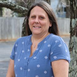 Photo of Amy Ludwick outside in front of a tree. She is wearing a blue shirt with a white spotted pattern. Amy has long brown hair. 